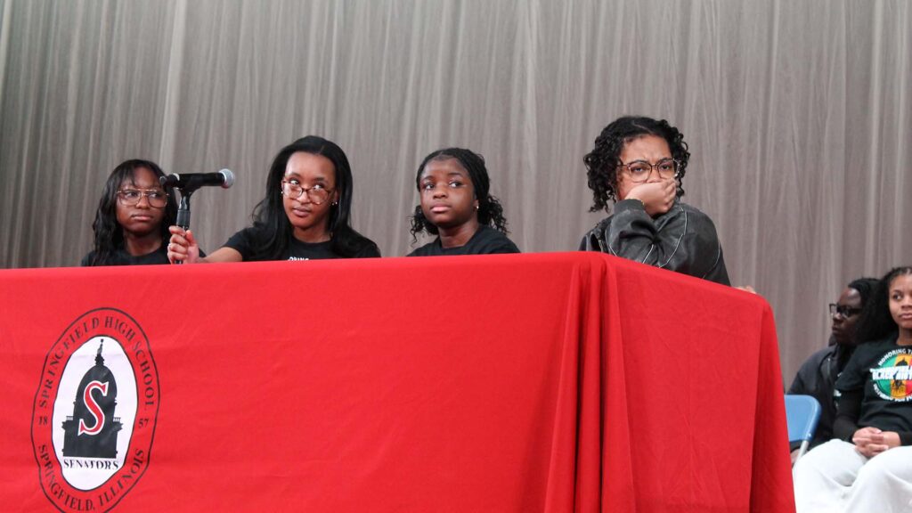 Four young women are seated at a table covered in a red table cloth with the Springfield High School emblem at the center. One looks over her glasses while holding the microphone stand. Another contemplates with a hand over their mouth.