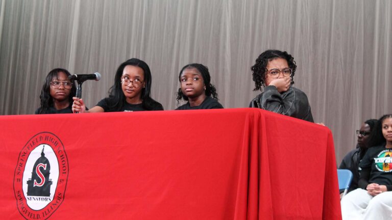 Four young women are seated at a table covered in a red table cloth with the Springfield High School emblem at the center. One looks over her glasses while holding the microphone stand. Another contemplates with a hand over their mouth.