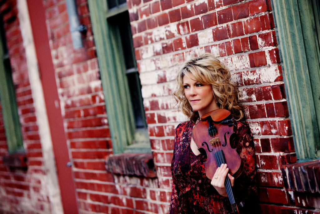 A woman holds a fiddle while posing in front of a brick building.