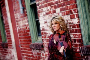 A woman holds a fiddle while posing in front of a brick building.
