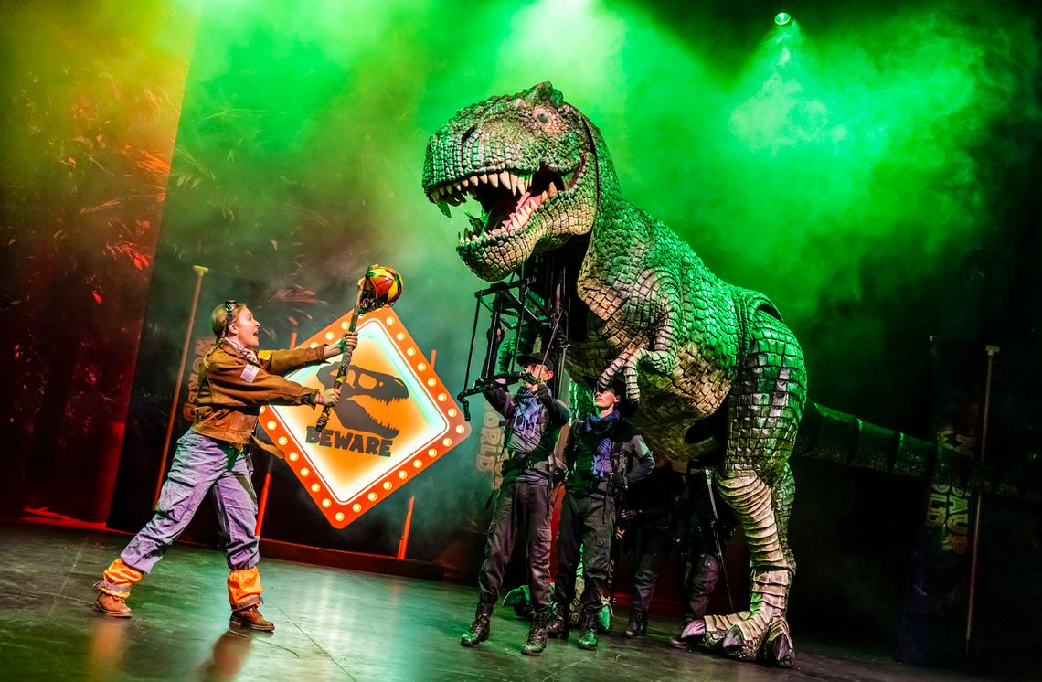 A person holds a stick-like object toward a large dinosaur puppet.