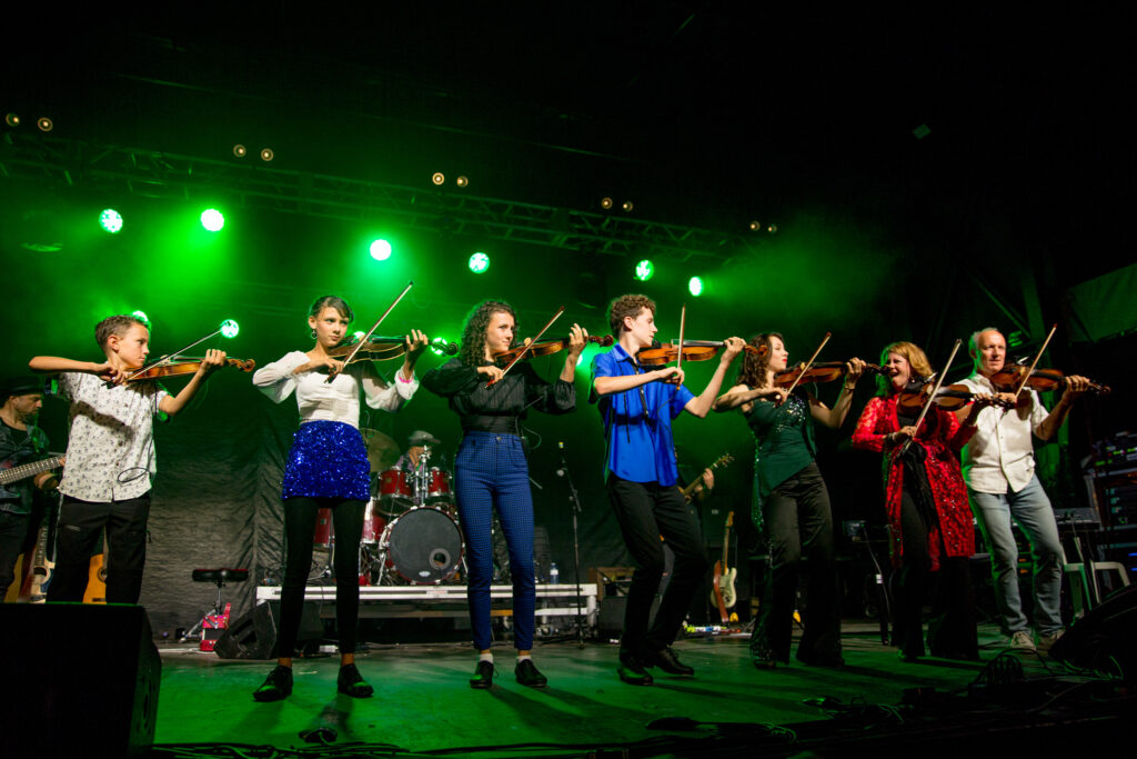 Courtesy Natalie MacMaster and MacIntyre Purcell Publishing Inc. Seven performers, including children and adults, play instruments on a stage.