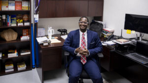 A man wearing a suit smiles while sitting at a desk chair in his office. Books about public health and leadership fill a tall bookshelf next to the desk.