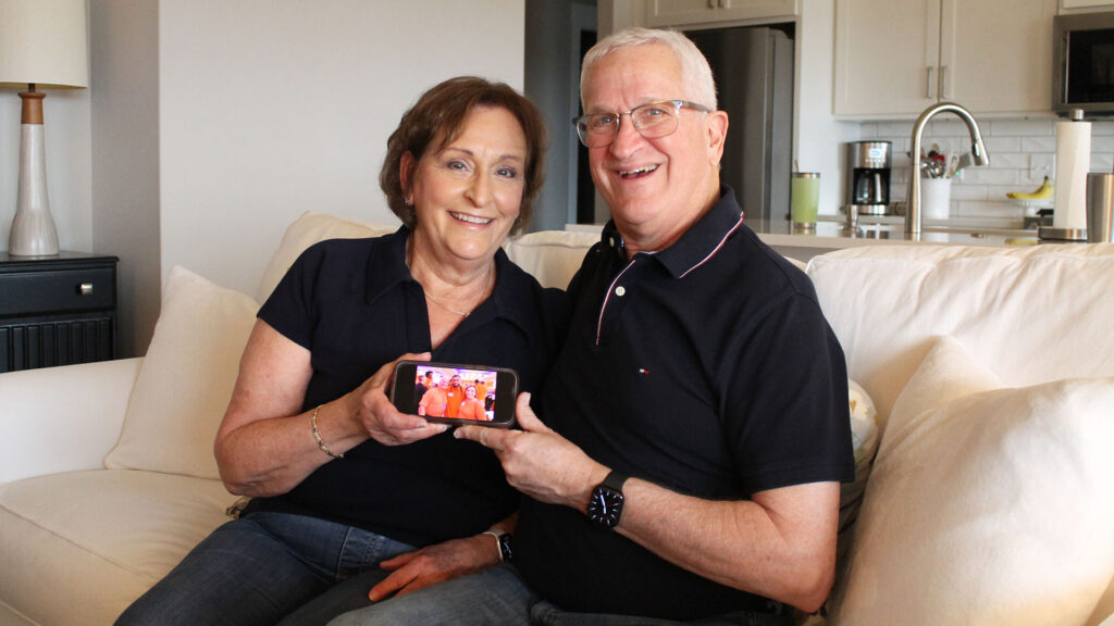 A middle-aged white couple grins while holding a phone together. The phone shows a recent picture of them with a basketball player.