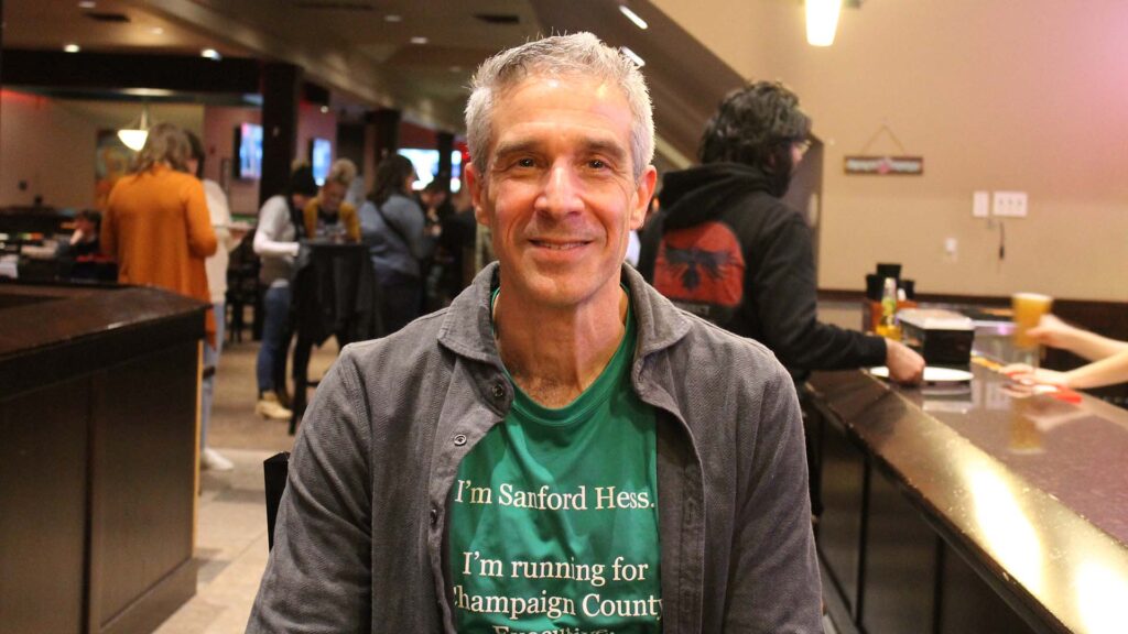 A white man with silver hair and a green t-shirt that says "I'm Sanford Hess. I'm running for Champaig County Executive." He's sitting at a bar with people in the background of the bar behind him.