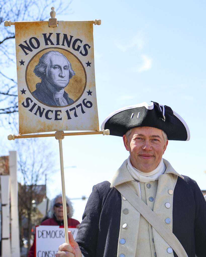A person dressed in a costume next to a sign that reads "No Kings Since 1776"