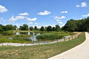 A sidewalk in a park with grass, a small body of water, trees and the sky