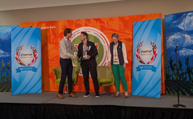 C16 Biosciences cofounder David Heller (center) holds a silver trophy while shaking hands with a man. A woman stands to his right on a stage in front of signs that say Startup World Cup.
