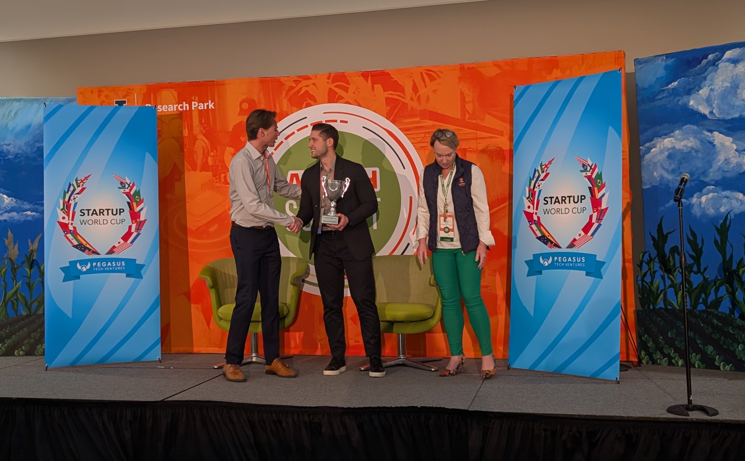C16 Biosciences cofounder David Heller (center) holds a silver trophy while shaking hands with a man. A woman stands to his right on a stage in front of signs that say Startup World Cup.