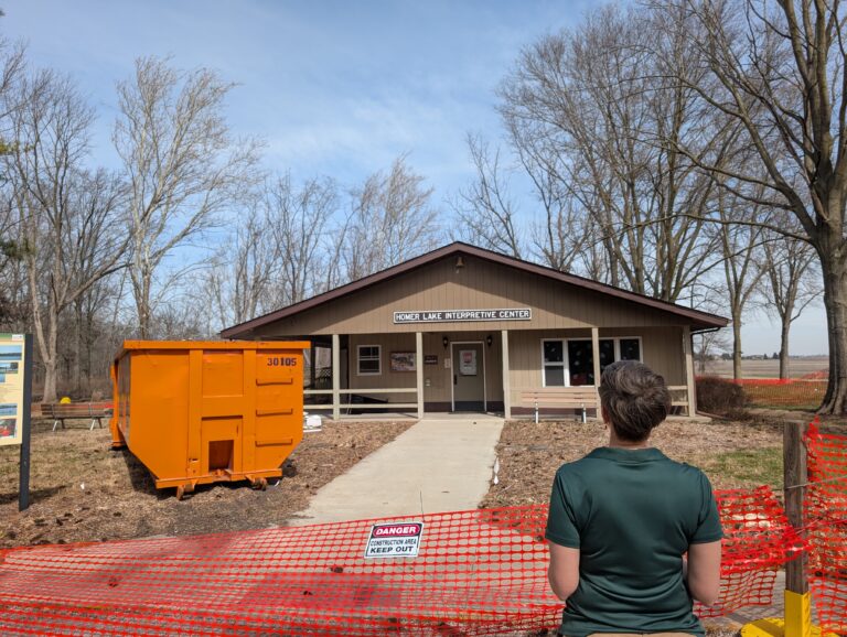 Champaign County Forest Preserve District Executive Director Lorrie Pearson looks onto the Homer Lake Interpretive Center, which is surrounded by orange fencing and has a yellow dumpster in front of it.