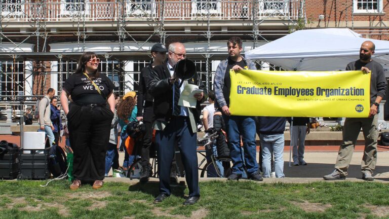 A man speaks into a megaphone in front of a university building. Two people hold a yellow sign that says "Graduate Employees Organization." The protest took place on the University of Illinois Urbana-Champaign campus Wednesday, April 8, 2026.