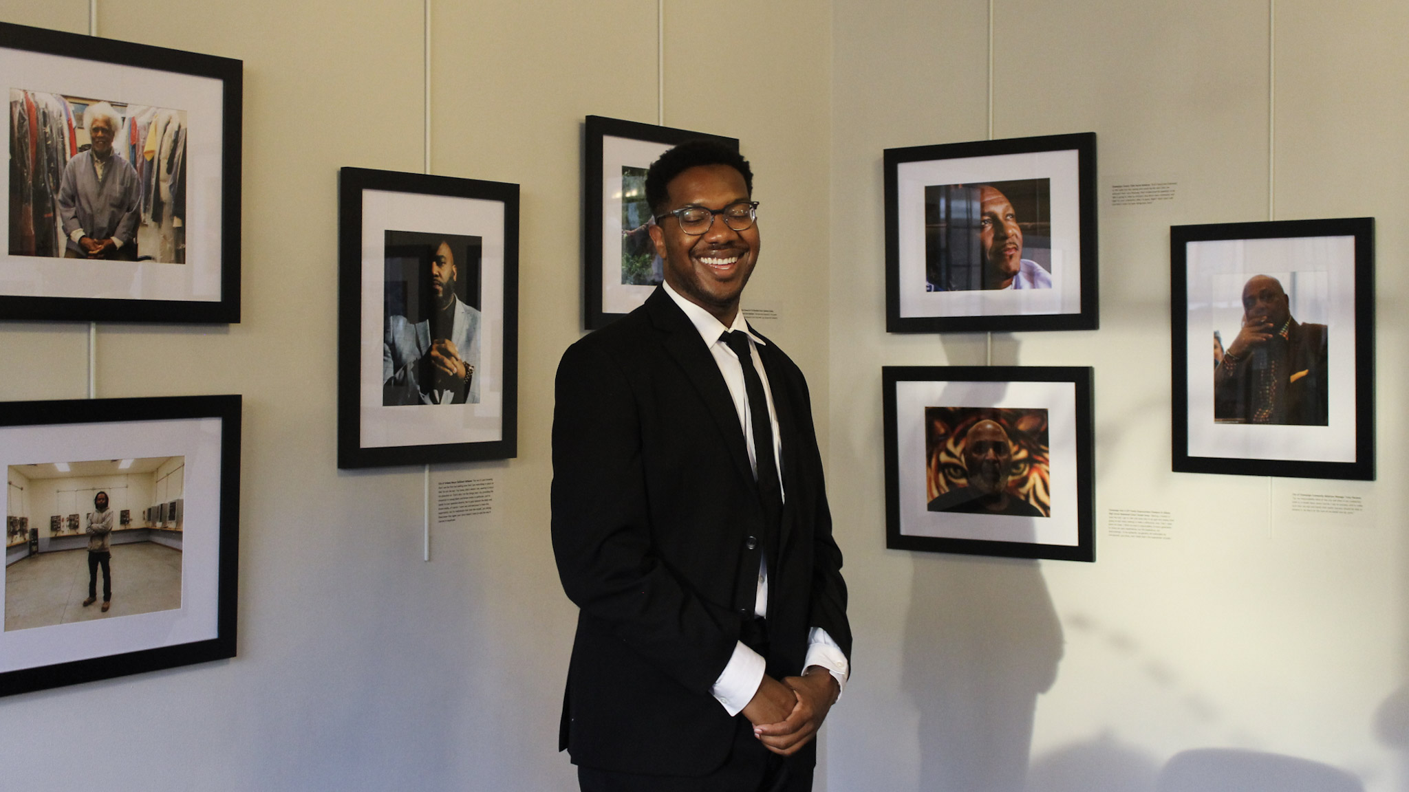 A laughing, young Black man stands in the corner of an art gallery of framed photos.