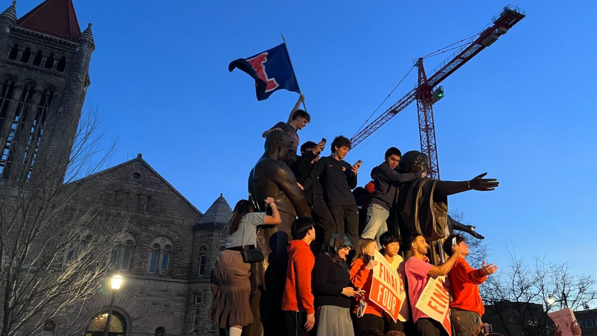 A group of people climb a bronze statue at dusk. Many people hold signs that say, "Final Four."