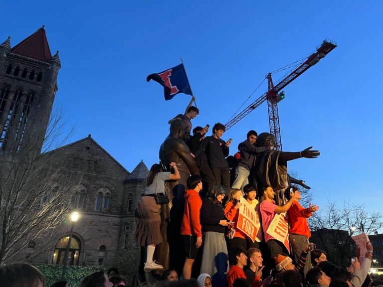A group of people climb a bronze statue at dusk. Many people hold signs that say, "Final Four."