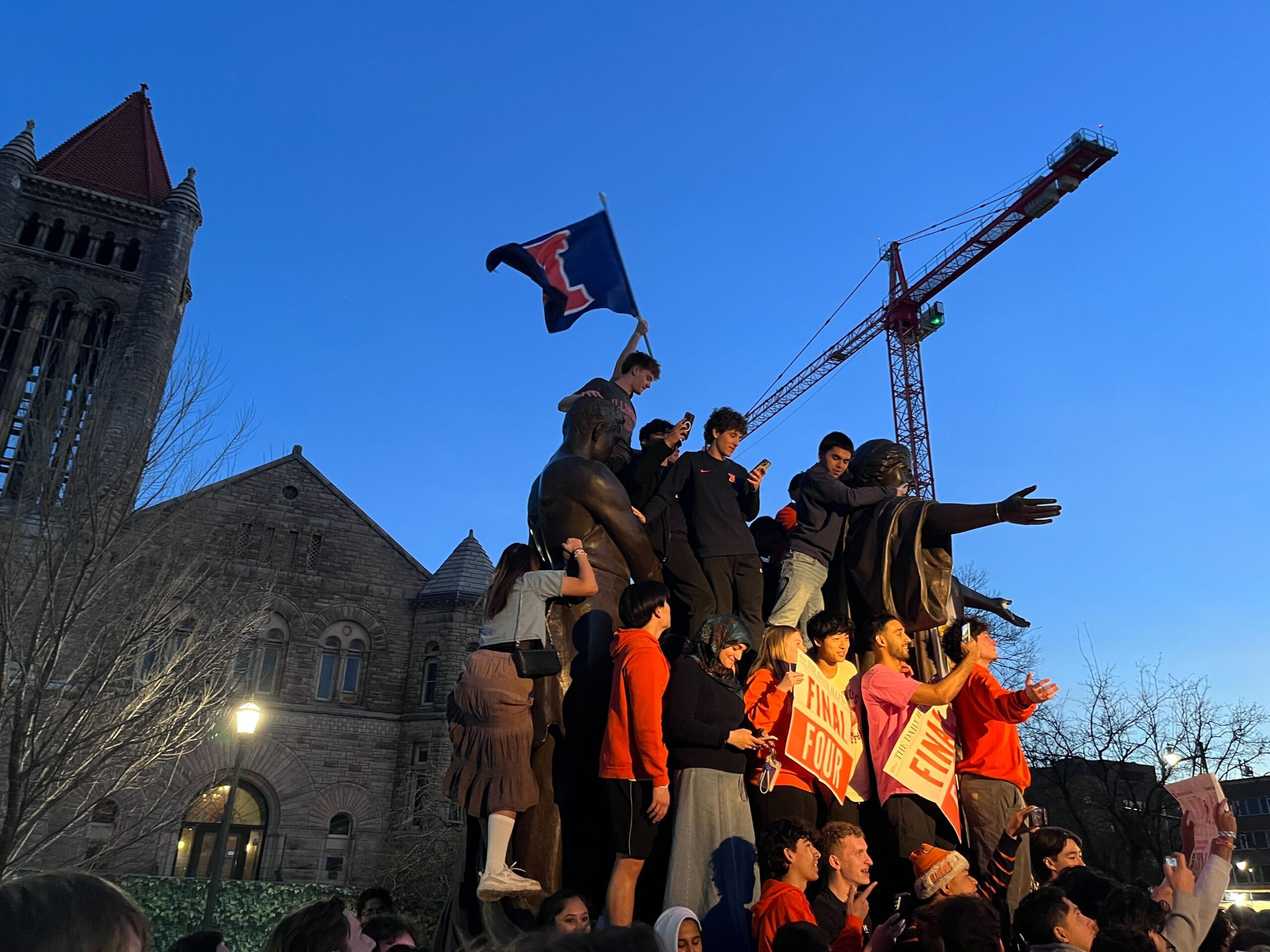 A group of people climb a bronze statue at dusk. Many people hold signs that say, "Final Four."
