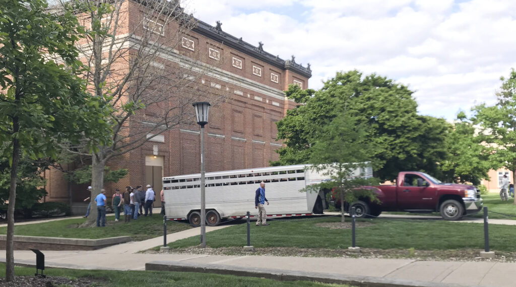 Several peoples stand outside of a long, silver trailer hitched to a red truck parked behind a brick campus building.