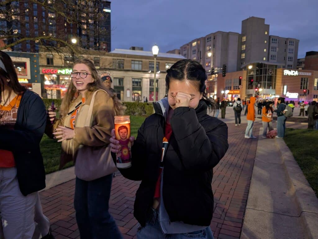 A woman places her right hand over her face while holding a lit candle with Michael Cera depicted as a saint in her left hand.