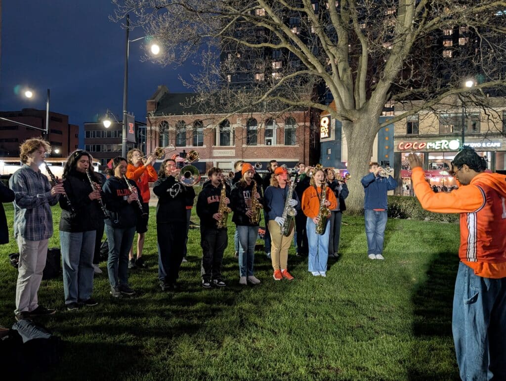 Members of the University of Illinois marching band play their instruments while their conductor stands in front of them.