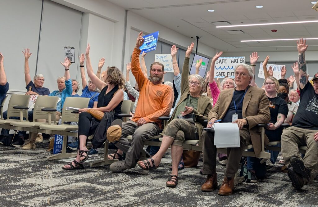 People sitting in chairs raise their hands. Some hold signs in support of a data center moratorium.