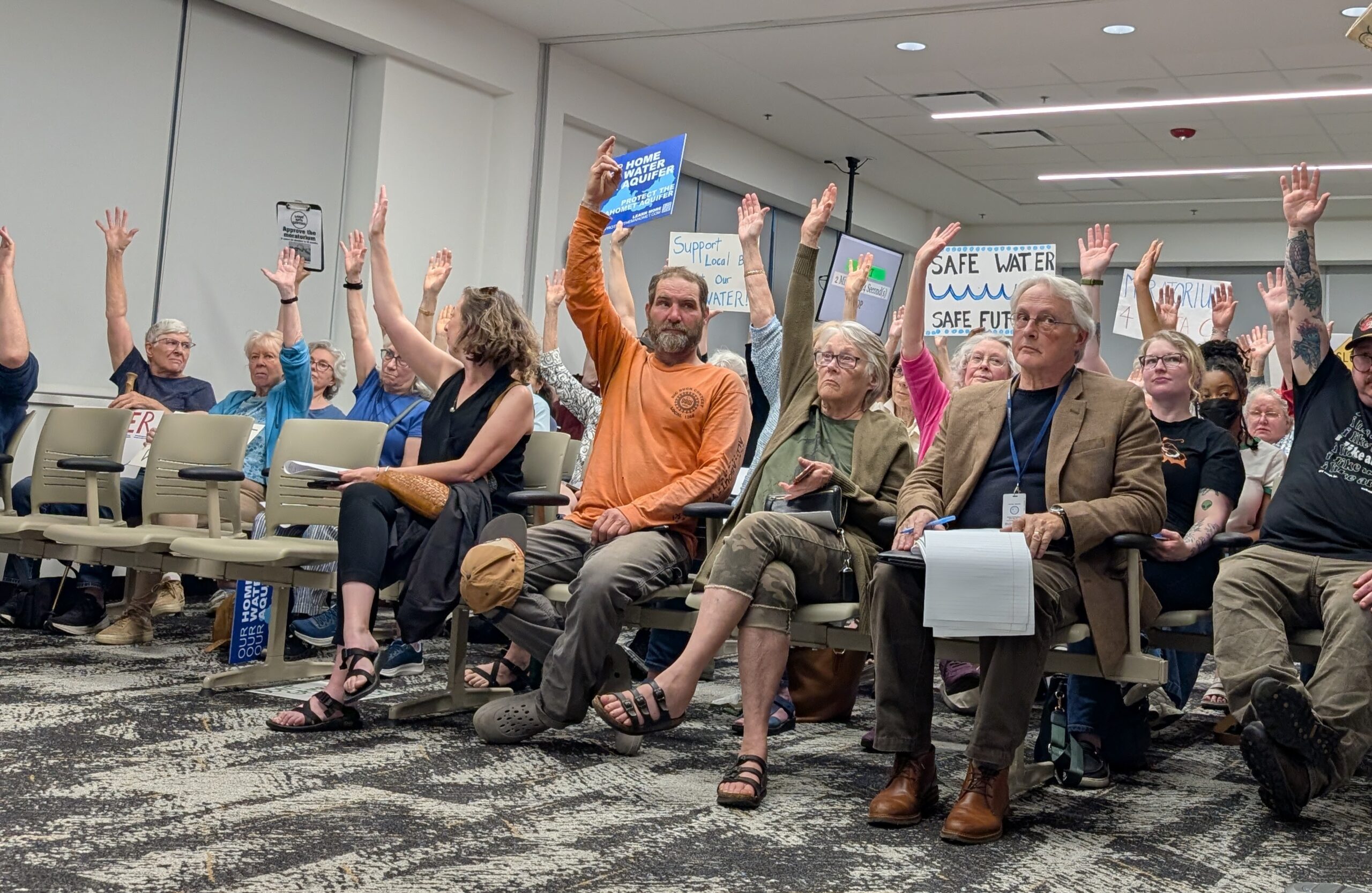 People sitting in chairs raise their hands. Some hold signs in support of a data center moratorium.