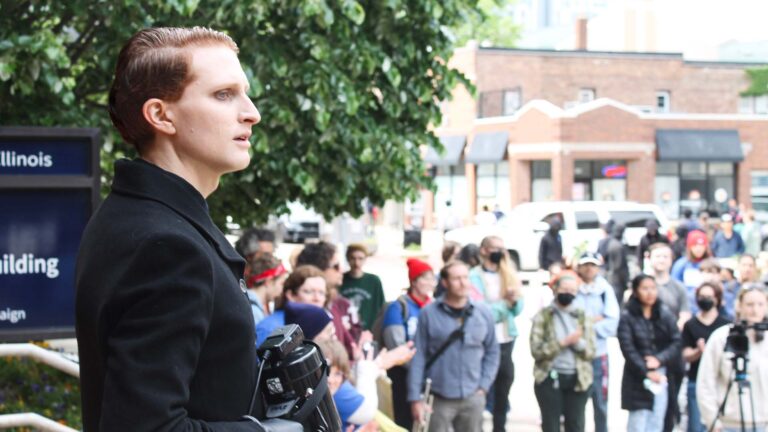 A person with short red hair and a black jacket holds a megaphone. In the background are protesters and a camera person.