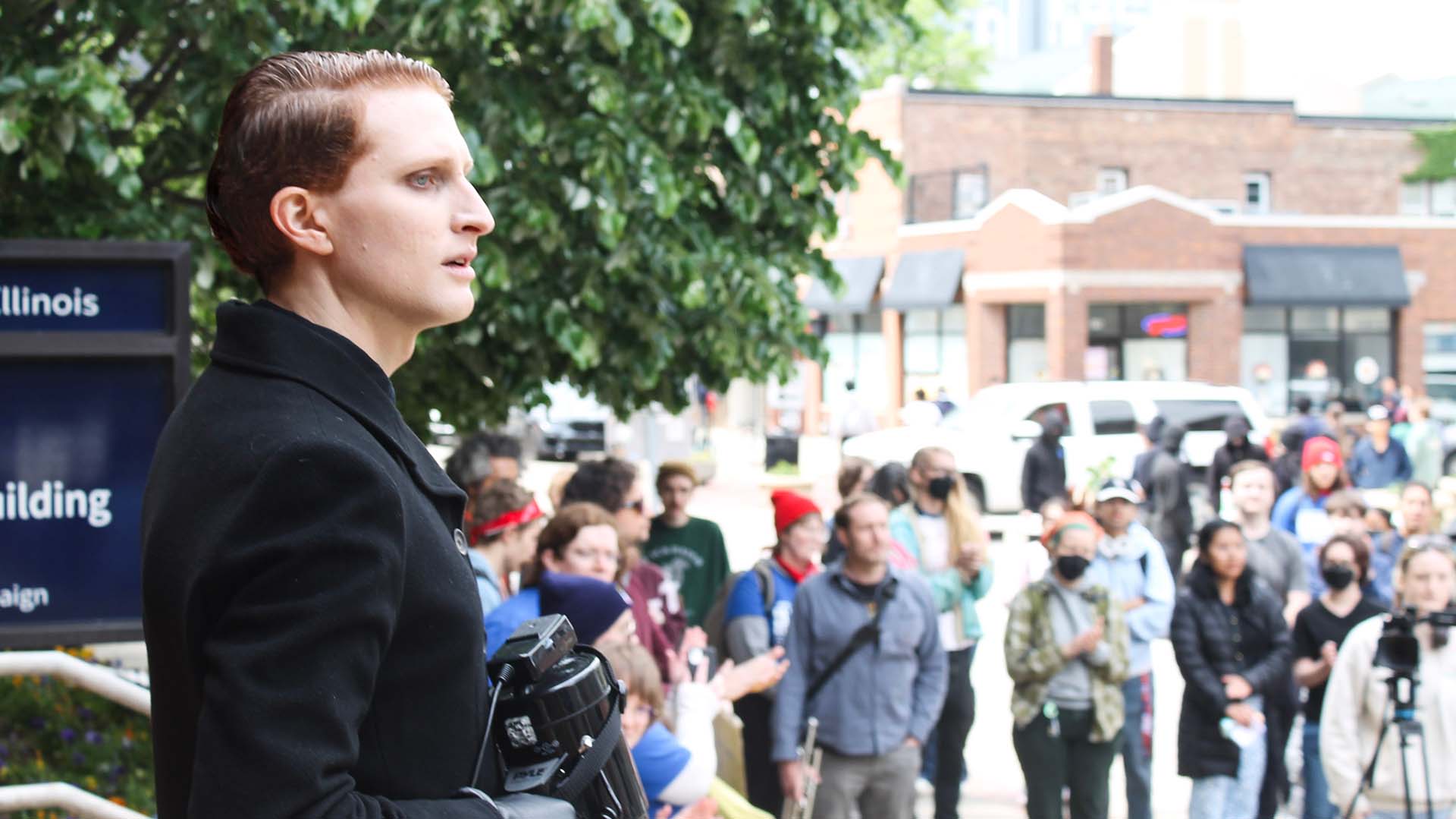 A person with short red hair and a black jacket holds a megaphone. In the background are protesters and a camera person.