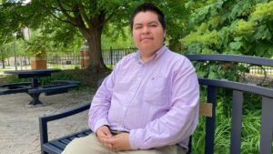 A young man in a lilac button-down sits on a bench outside Illinois Public Media's station on the University of Illinois Urbana-Champaign campus.