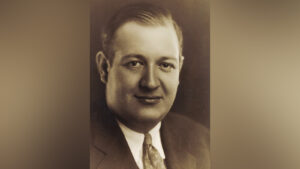 A black and white portrait of a white man in a suit, slightly smiling at the camera.