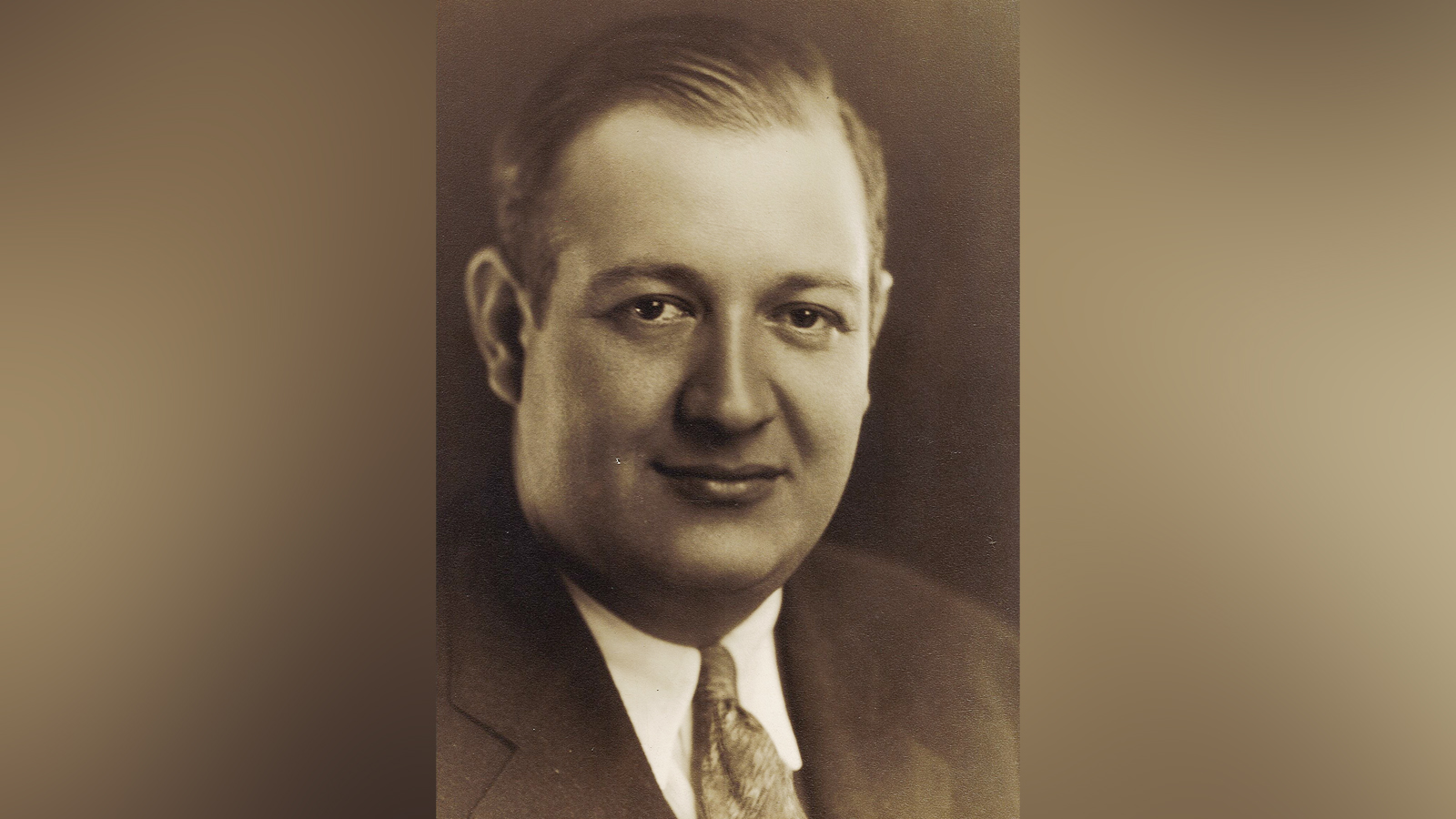A black and white portrait of a white man in a suit, slightly smiling at the camera.