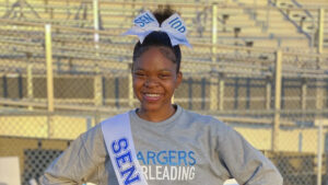 A Black teen girl stands in front of bleachers and smiles at the camera, with her hair up in a bun, a large bow in her hair and a sash that says "Senior" over her shoulder. She wears a grey shirt that says "Chargers Cheerleading."