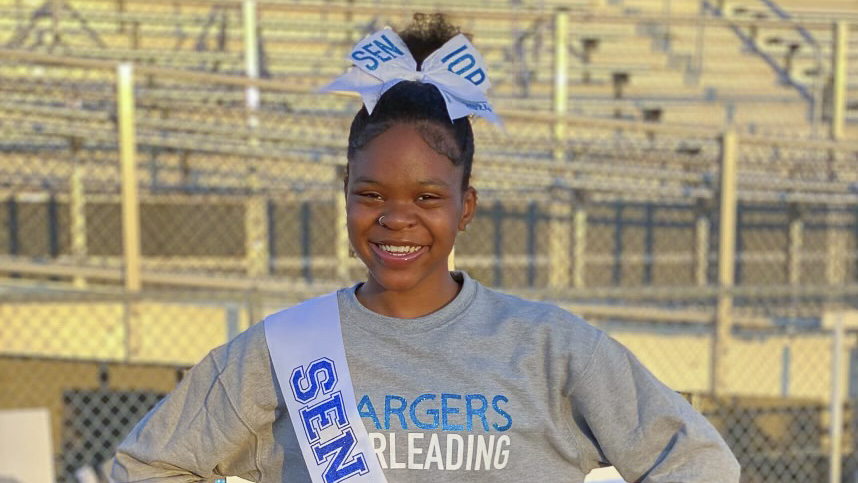 A Black teen girl stands in front of bleachers and smiles at the camera, with her hair up in a bun, a large bow in her hair and a sash that says "Senior" over her shoulder. She wears a grey shirt that says "Chargers Cheerleading."
