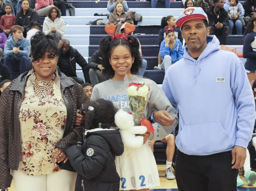 A Black teen girl holds flowers and stands between a man and a woman, whose arms are linked with her. A younger Black girl stands in front of her and holds onto the woman's arm. They stand in front of indoor gymnasium bleachers and smile at the camera.