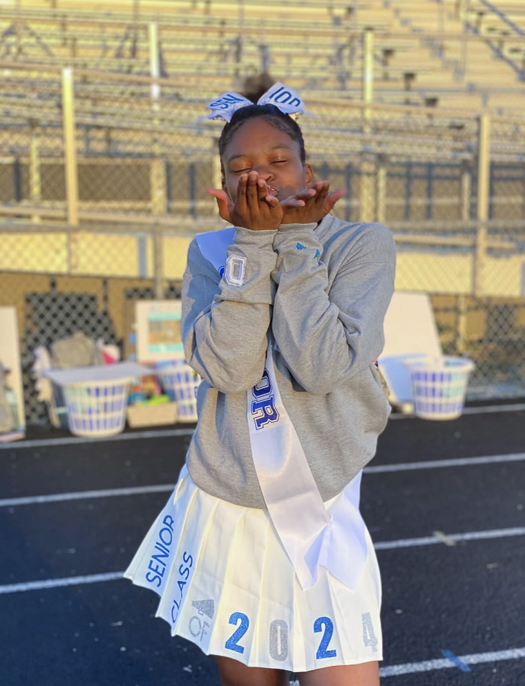 A Black teen girl blows a kiss at the camera while standing in front of bleachers. She wears her hair in a bun with a large bow, a grey shirt, a white cheerleading skirt, and a white sash over her shoulder.
