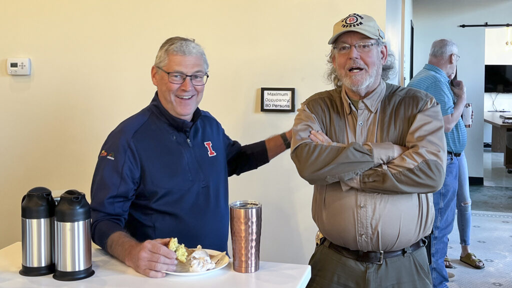 Two older white men stand together. One man wears a blue quarter zip with an orange "I" and is resting his hand on the shoulder of the other man, who wears a khaki baseball cap with a Pour Bros. Taproom logo and a tan button-up shirt.