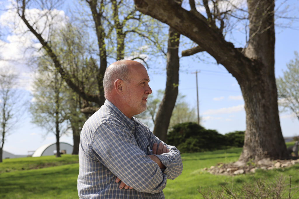 A man stands outside in a field, with his arms folded
