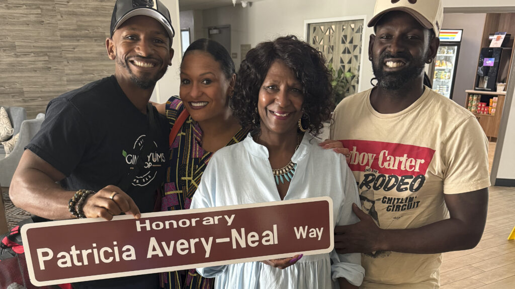 A woman and her three adult children stand and smile at the camera, holding a brown street sign that reads "Honorary Patricia Avery-Neal Way."