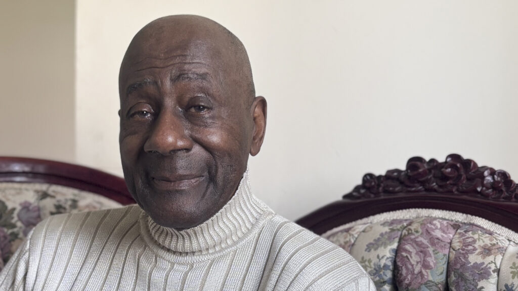 An older Black man wearing a cream-colored turtleneck sweater sits on an antique sofa.