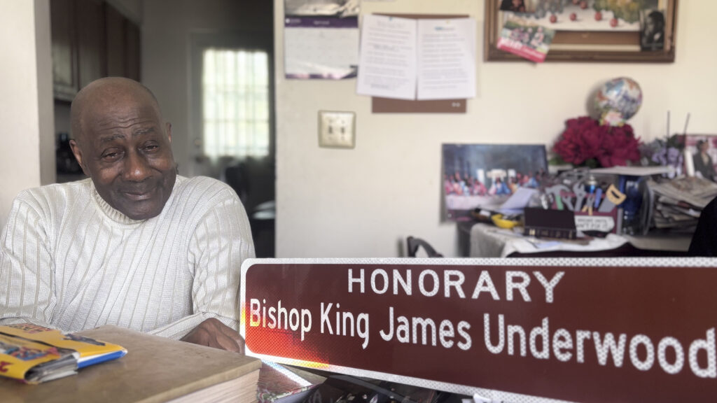 An older Black man wearing a cream-colored turtleneck sweater sits at a desk and holds a brown street sign reading "Honorary Bishop King James Underwood Way."