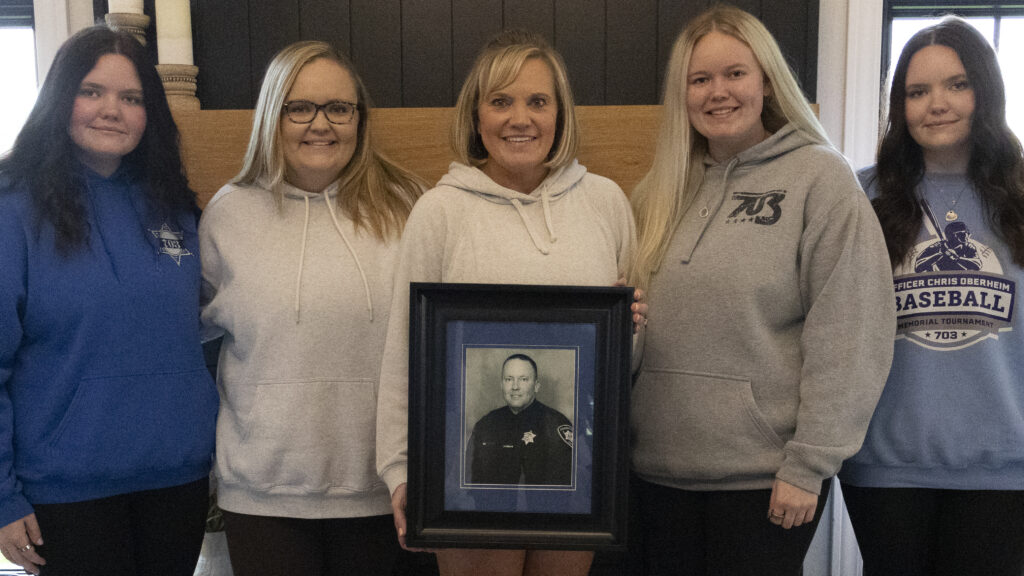 Five white women stand in line, the woman in the center holds a black and white framed photo of a man in a police uniform.