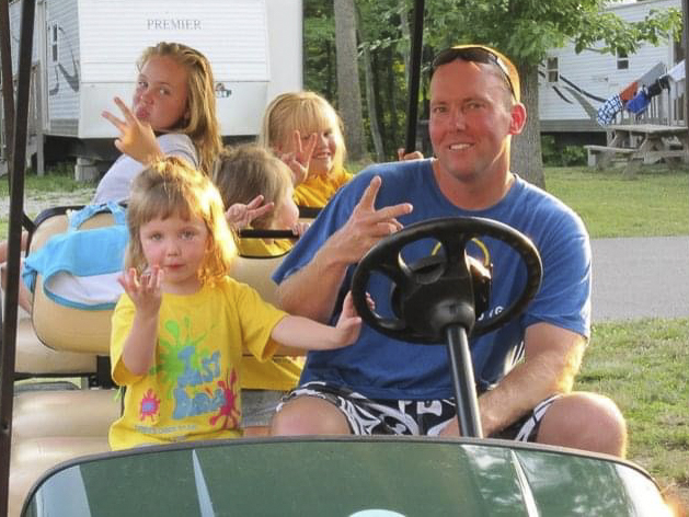 A man and four young girls sit in a golf cart, smiling and flashing peace signs at the camera.