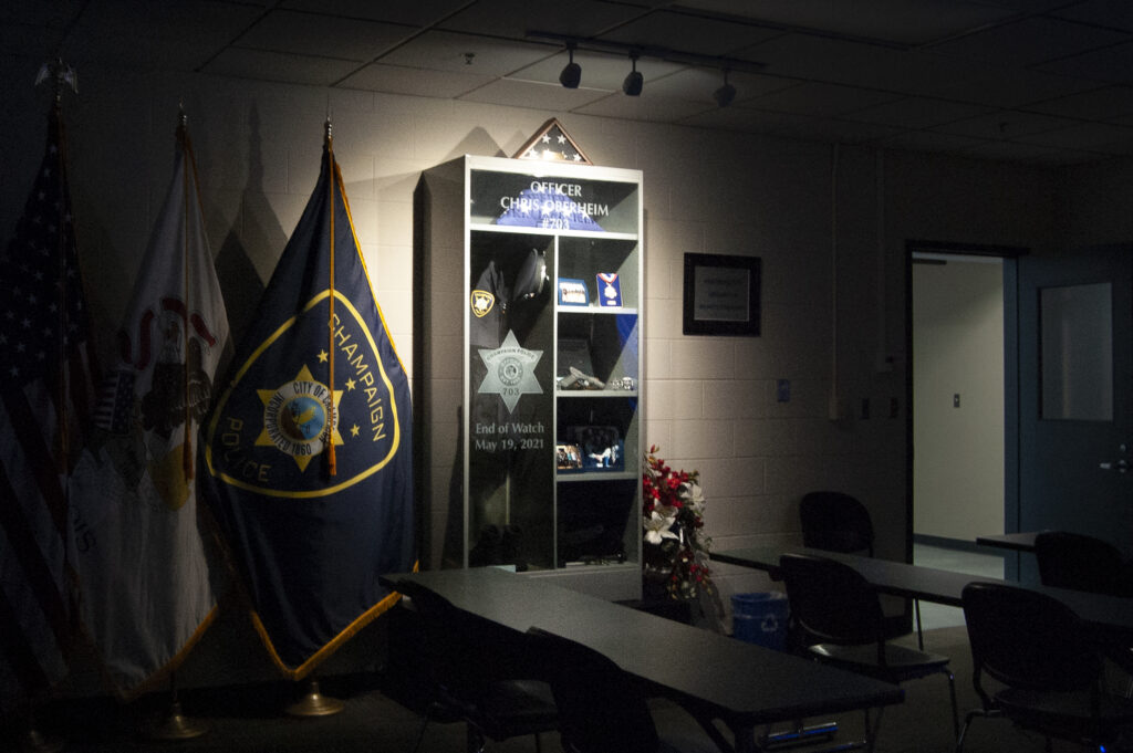 A dark meeting room with a single light illuminating a shelf holding a gun, a uniform and other items belonging to the fallen police officer Chris Oberheim.