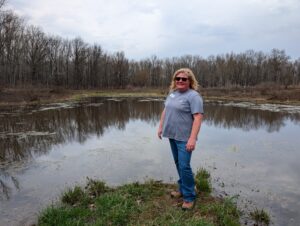 East Central Illinois FrogWatch chapter coordinator Morgan Duerksen-Balk stands on a patch of grass in front of a wetland wearing sunglasses. There's a treeline behind her.
