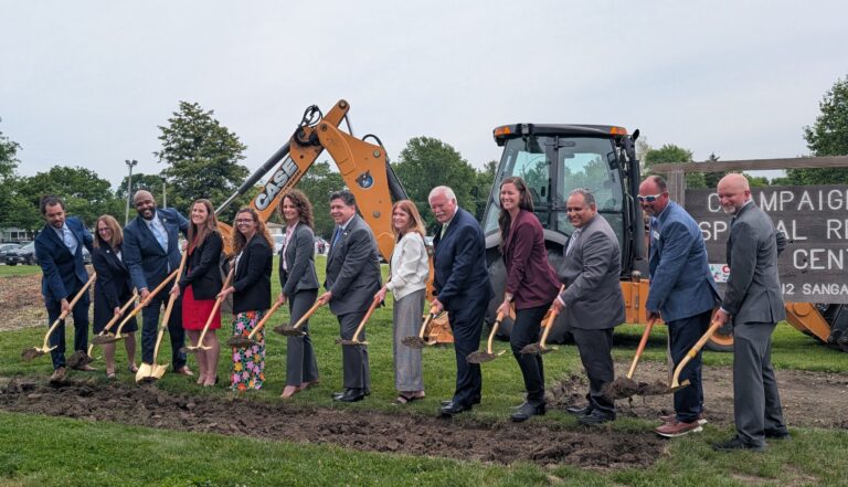 Officials hold shovels filled with dirt in front of a piece of construction equipment.