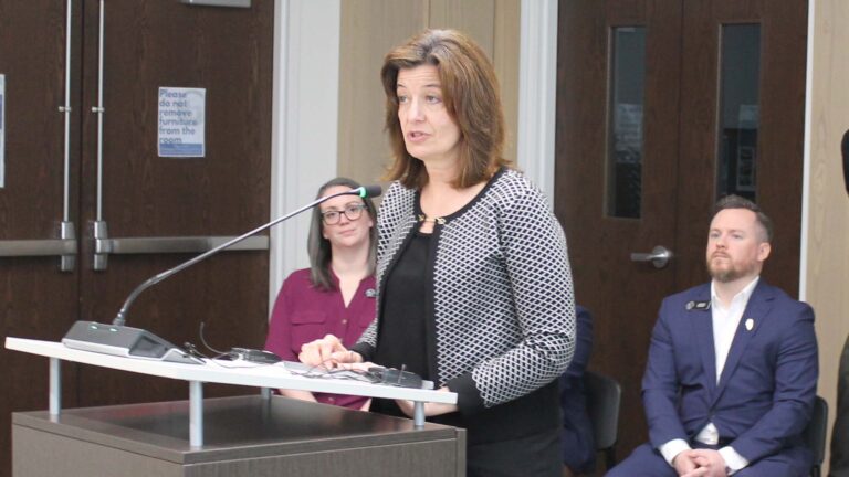 A woman with brown hair stands at a podium with a concerned expression.