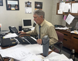Gary Scott sits at his desk in the WLDS-WEAI office, with stacks of papers across his workspace.