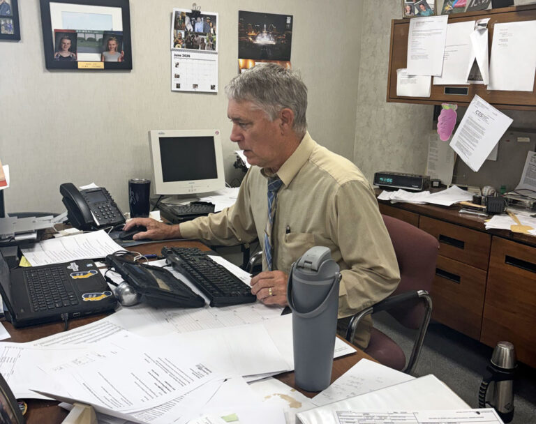 Gary Scott sits at his desk in the WLDS-WEAI office, with stacks of papers across his workspace.