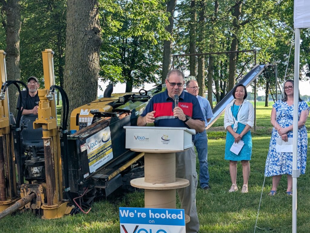 A man speaks into a microphone while standing behind a podium. To his left sits a directional boring machine.