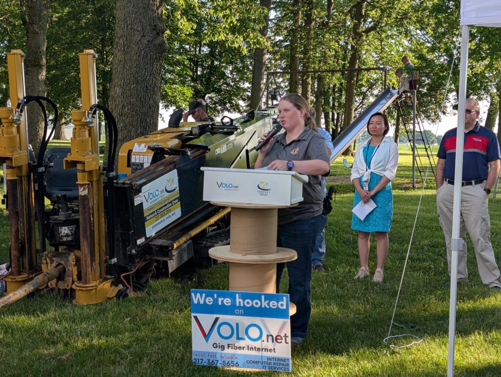 A woman speaks into a microphone behind a podium. In front of the podium is a sign that reads, "We're hooked on Volo.net gig fiber internet." To her left is a directional boring machine.