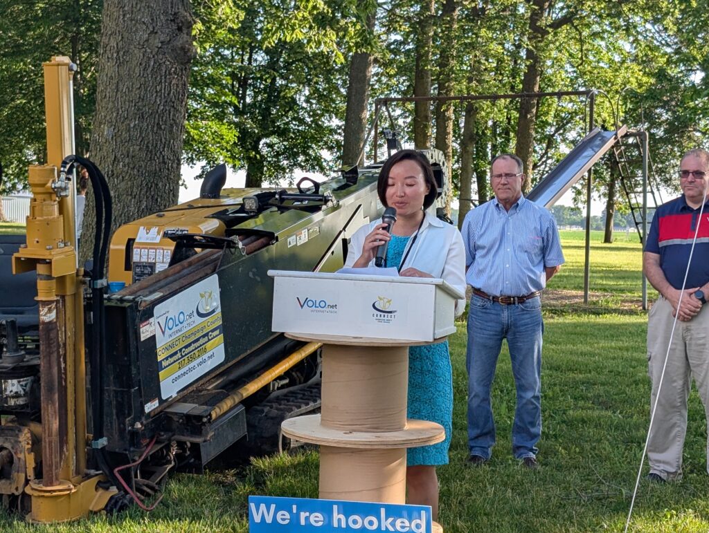 A woman speaks into a microphone behind a podium. A directional boring machine sits to her left.