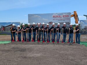People in matching black polo shirts, jeans and white construction helmets hold red shovels stuck in a mound of dirt in front of a rendering of the new Rural King campus.